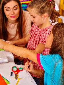 Close up of children girl with young female teacher are making something out of colored paper on table in primary school. Children craft lesson in primary school. Teacher learn children.